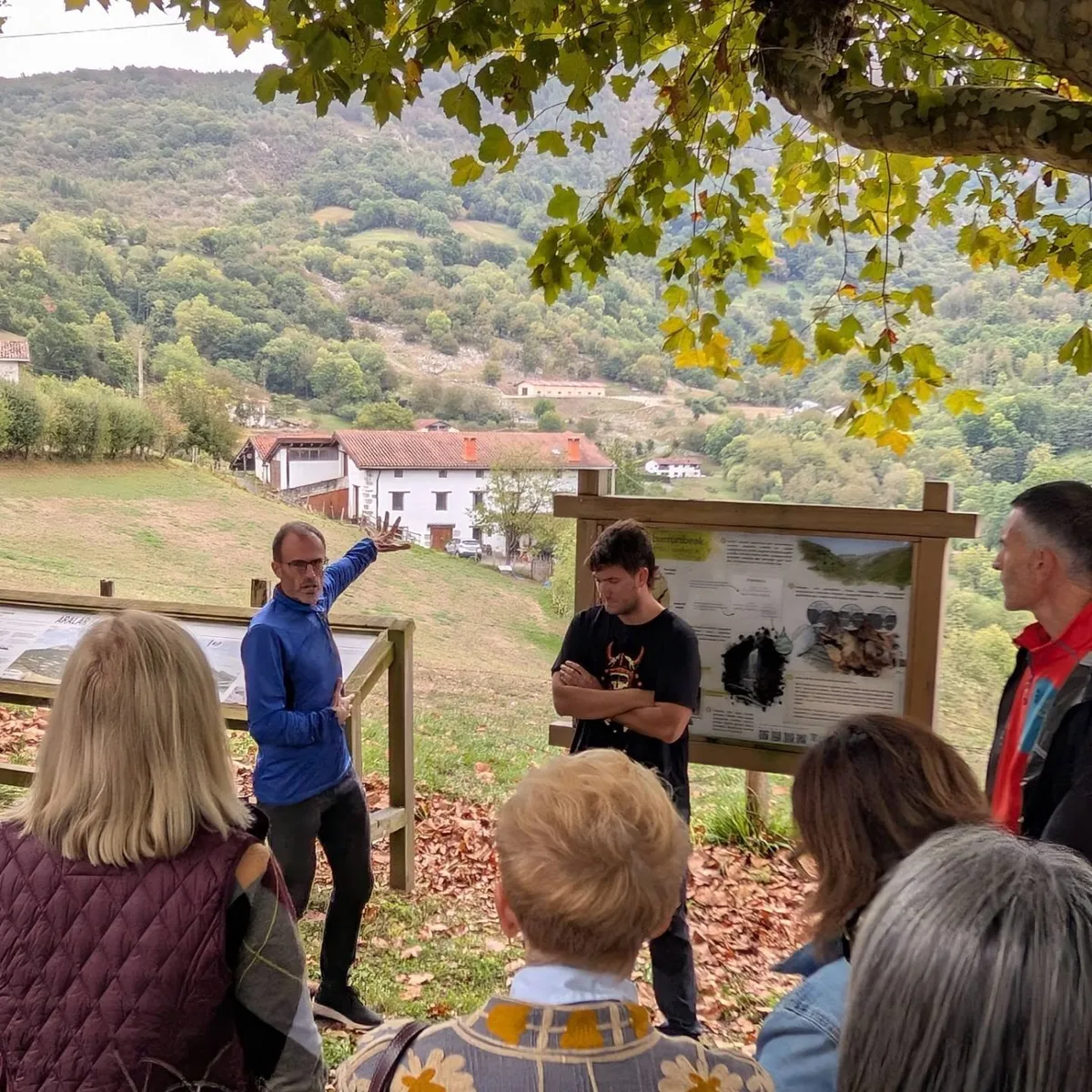 Un guía explica el paisaje junto a paneles informativos ante el grupo, con caseríos y montes al fondo